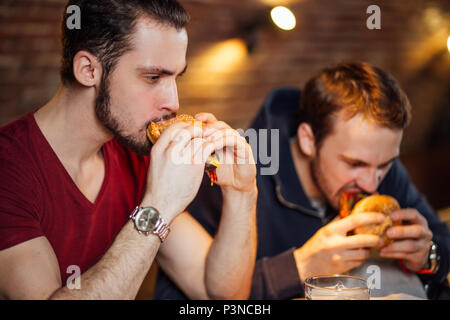 Two happy male friends eating tasty burgers in bar. Stock Photo