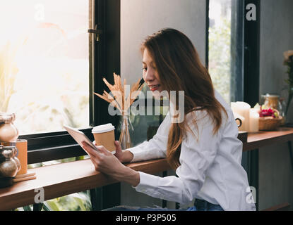 Asian Businesswoman holding coffee cup and reading report on tablet computer on wood stool table at coffee shop near window,woking outside office life Stock Photo