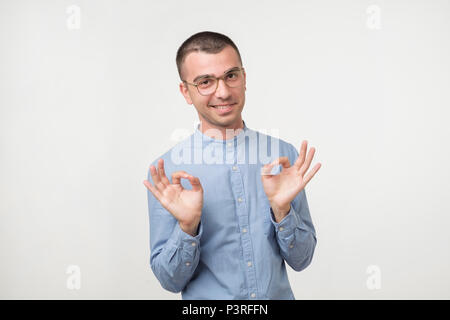 A young latin handsome man in blue shirt isolated on gray background showing ok sign. Concept of success, triumph at studies or job project Stock Photo
