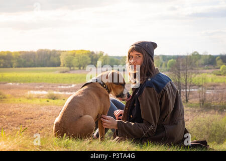 Beautiful hiking girl sit on hill with pet dog at walk. Young female person together with staffordshire terrier at meadow on warm sunny afternoon Stock Photo