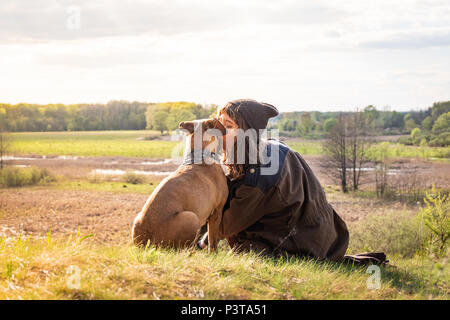 Beautiful hiking girl sit on hill and kiss pet dog at walk. Young female person together with staffordshire terrier at meadow on warm sunny afternoon Stock Photo
