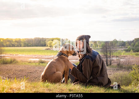 Beautiful hiking girl sit on hill with pet dog at walk. Young female person together with staffordshire terrier at meadow on warm sunny afternoon Stock Photo