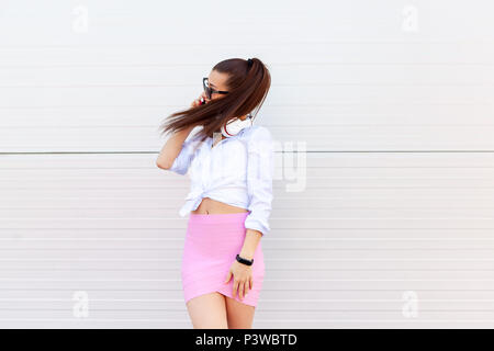 Fashion portrait of pretty smiling woman in sunglasses talking on the smartphone against the grey wall. Wireless Headphones on the neck, fitness brace Stock Photo