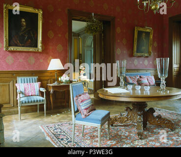 Blue upholstered chairs at antique circular table in French country dining room with red wallpaper and antique silk rug Stock Photo