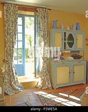 Blue+white floral curtains on blue French windows in country dining room with blue+cream wooden dresser Stock Photo