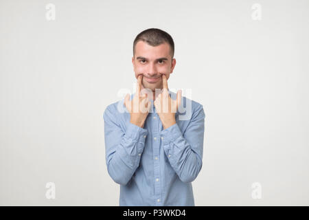 Handsome young man in blue shirt trying to smile instead of bad depressed mood. Making faces. Negative facial emotion Stock Photo