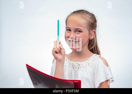 A cute 10-year-old caucasian schoolgirl smiling while holding up a pencil in the air and her note pad on her knee against a light background Stock Photo