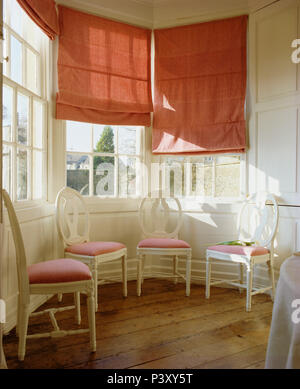 Pink blinds on bay window above set of white painted spoon back chairs with pink seats in dining room with wooden flooring Stock Photo