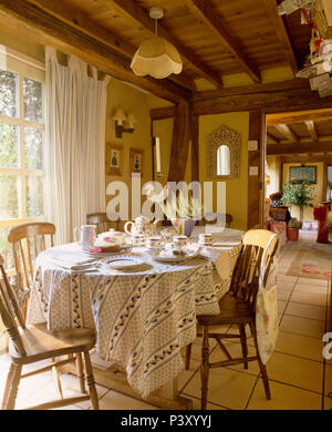 Blue+white cotton cloth on table set for tea in cottage dining room with old wooden chairs Stock Photo