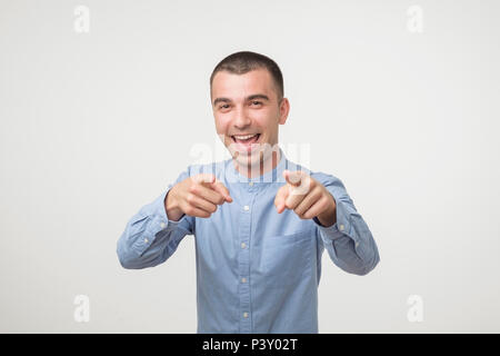 Indoor shot of cheerful young man in blue shirt indicates happily at you, chooses to compete, has positive expression. Handsome clever student makes a Stock Photo