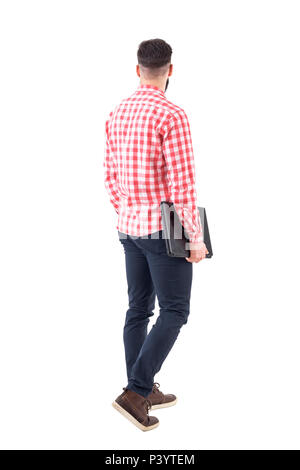 Rear view of young adult business man carrying laptop under arm walking and looking away. Full body isolated on white background. Stock Photo