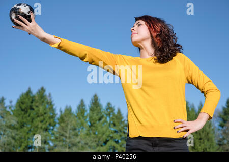 The whole world in her hand: Young woman in stylish yellow pullover holding earth in her hand in front of sky and trees. Save the earth concept. Stock Photo