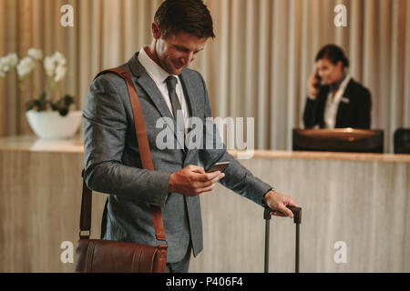 Happy businessman in hotel lobby using cellphone. Male guest arriving at his hotel with phone and suitcase. Stock Photo