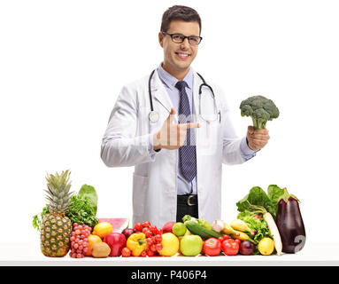 Doctor with broccoli pointing behind a table with fruit and vegetables isolated on white background Stock Photo