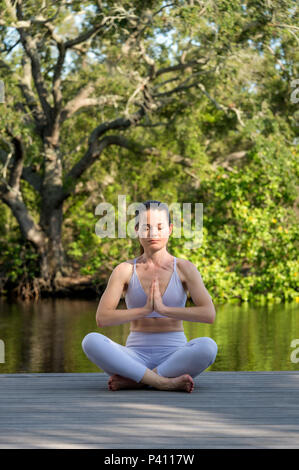 woman sitting on deck by water meditating and practicing yoga Stock Photo