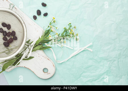 Mulberry in a colander and daisies on a kitchen board on a turquoise background Stock Photo