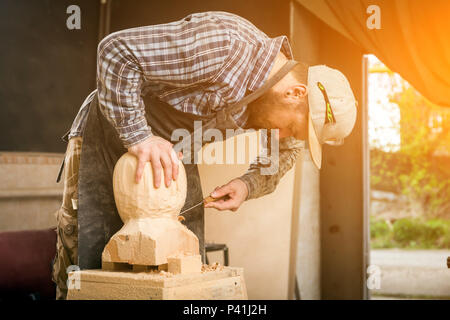 Young man carpenter in a work clothes  saws a man's head with a tree , using a chisel  in the workshop, around a lot of tools for work Stock Photo