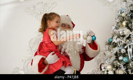 Santa Claus helping little girl to hang baubles on top of the Christmas tree Stock Photo