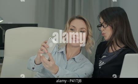Two female colleagues taking selfies with phone sitting at the desk Stock Photo