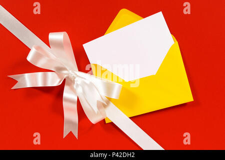 Christmas card with diagonal gift ribbon and bow in white satin on red paper background with yellow envelope and blank card Stock Photo