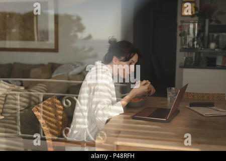 Young woman eating breakfast while looking into laptop in living room Stock Photo