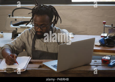 Carpenter writing in clipboard while using laptop Stock Photo