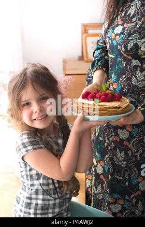 Mom and the girl are holding a plate with homemade pancakes and berries. Delicious breakfast at home. A happy family. Good morning. Stock Photo