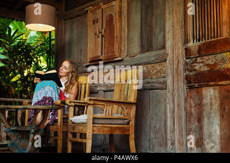 Attractive girl sit on open-air veranda of wooden bungalow with tropical garden view, read romance in paper book. Young woman relaxing in luxury villa Stock Photo