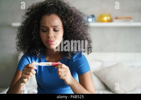 Disappointed hispanic girl getting unexpected result from pregnancy test kit. Sad young latina woman sitting alone on her bed. Stock Photo
