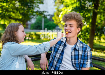 Young couple in love. Summer in park sit on a bench. A girl is feeding a guy with food. The guy smiles happily. The concept of a happy relationship. Stock Photo