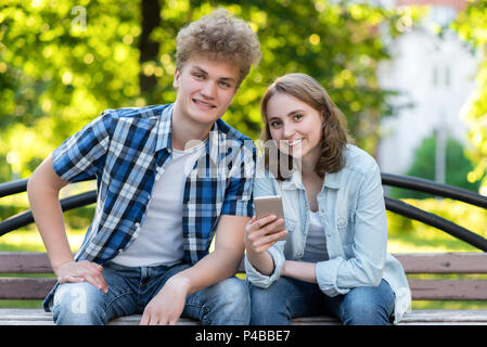 Young couple girl boy. Summer in park on a bench. In his hands holds a smartphone. Happy smiling close-up. Emotions of happiness and pleasure. The student rests after school. Stock Photo