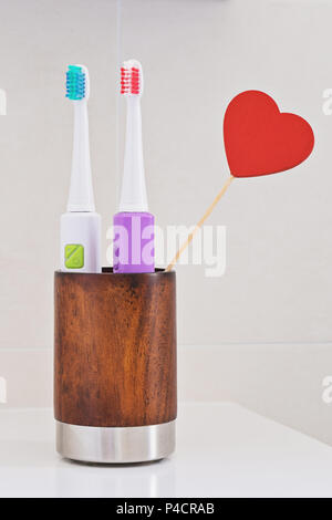 Two electric toothbrushes in wooden glass on sink in the bathroom with red heart. Personal hygiene like concept. Greeting card to celebrate Valentine' Stock Photo