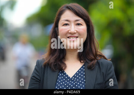 Mature happy beautiful Asian businesswoman smiling in the streets outdoors Stock Photo