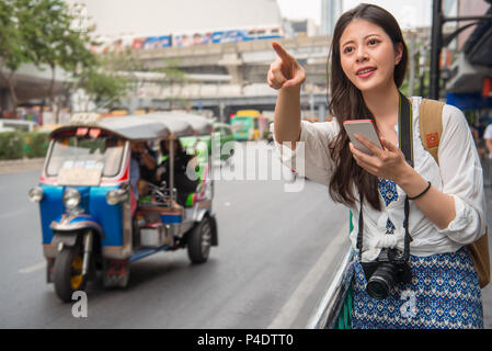 Asian woman traveller standing beside the street and pointing to the front to look for a cab with her cellphone. Stock Photo