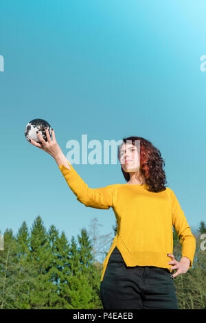 The whole world in her hand: Young woman in stylish yellow pullover holding earth in her hand in front of sky and trees. Save the earth concept. Stock Photo