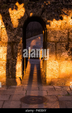 Man sized entry way through the medieval city walls of Trancoso, Portugal. With shadow of person and tree being cast onto the sunset lit wall and path Stock Photo