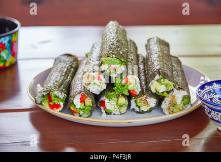 Long sushi rolls with rice, nori and vegetables lying on ceramic plate standing on wooden table with kitchenware with traditional rustic ornaments Stock Photo