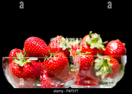 Close up of two crystal glass dessert dishes full of fresh juicy strawberries with leaves & stalks, black background. Fruit bowl isolated indoors. Stock Photo