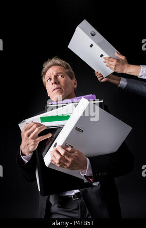 scared overworked mature businessman holding pile of folders isolated on black Stock Photo