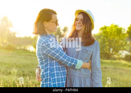 Parent and teenager, mother and 14 year old daughter embrace smiling in nature. Background sunset, rustic landscapes, green meadow Stock Photo