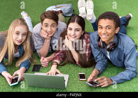 portrait of multicultural smiling teenagers using laptop together while resting on green grass Stock Photo
