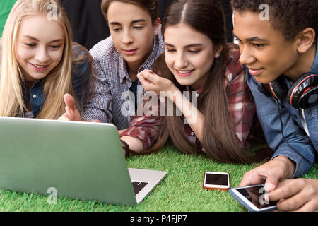 portrait of multicultural smiling teenagers using laptop together while resting on green grass Stock Photo
