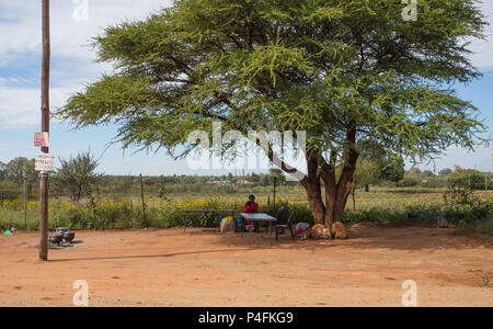 Rural scene of an African female and child sitting at an empty table under a Camel Thorn (Acacia) tree at the side of the road in South Africa Stock Photo