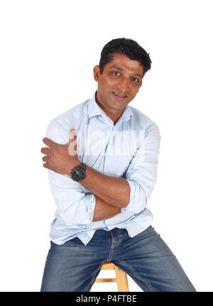 A handsome middle age East Indian man sitting on a bar chair in jeans and a blue shirt, isolated for white background Stock Photo
