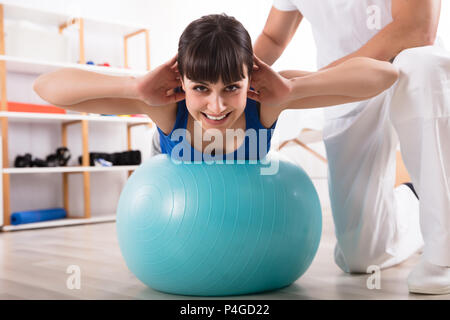 Physiotherapist Assisting Happy Woman While Doing Exercise On Fitness Ball Stock Photo