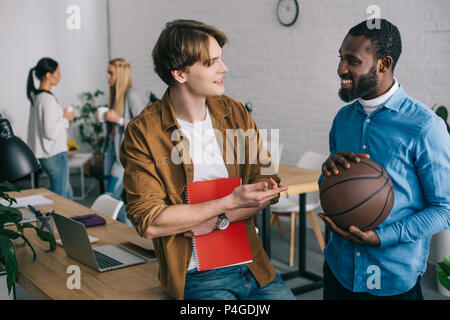 smiling african american businessman holding ball and talking to partner with textbook and two businesswomen standing behind with coffee