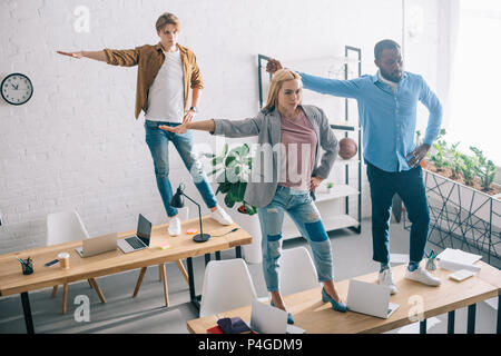 high angle view of happy multiethnic business colleagues having fun and dancing on tables in modern office Stock Photo