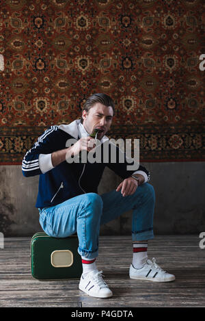 young man sitting on vintage little suitcase and trying to open beer with teeth in front of rug hanging on wall Stock Photo