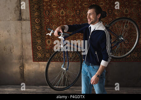 happy young man in vintage clothes carrying road bike on shoulder in front of rug hanging on wall Stock Photo