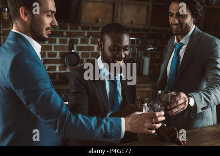 smiling young multiethnic male friends in suits clinking glasses of whiskey Stock Photo